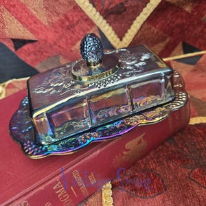 May include: A rectangular iridescent carnival glass butter dish with a matching lid. The lid features a textured pineapple-shaped finial and embossed grape vine patterns. The base has a scalloped edge with a matching embossed grape vine motif.