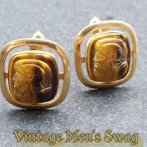 May include: A pair of gold cufflinks with a square design. Each cufflink features a brown tiger's eye stone with a carved image of a man's face.