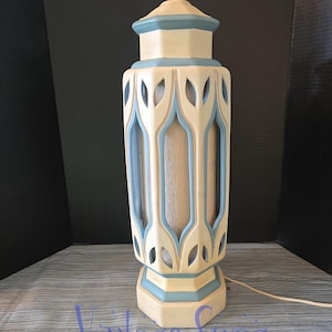 May include: A vintage table lamp with a cream-colored body and light blue accents. The lamp features a decorative cut-out design and a brass-colored lamp holder. The lamp has a cord and plug for electrical use.