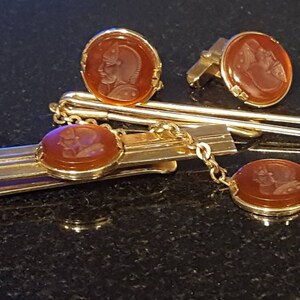 May include: A gold-toned tie bar, cufflinks, and chain set with brown cameo inlays. The cameos feature carved portraits.