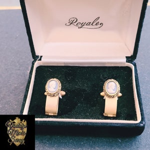 Op de afbeelding: Een paar gouden manchetknopen met een wit cameoportret in een zwarte fluwelen doos. De doos is gelabeld met "Royale."