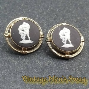 Könnte beinhalten: Ein Paar Manschettenknöpfe mit einem schwarzen und weißen Kameen-Design. Die Kamee zeigt eine weiße Silhouette von zwei Figuren, die auf einem schwarzen Hintergrund ringen. Die Manschettenknöpfe sind von einem goldfarbenen Metallrahmen umgeben.
