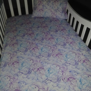 May include: A white crib sheet with a purple and blue watercolor print of Ariel from The Little Mermaid. The sheet is made of soft, breathable fabric and is perfect for a baby's crib.