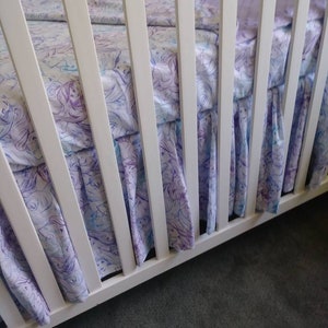 May include: White crib with a white and purple patterned fitted sheet and a white ruffled crib skirt. The crib skirt has a watercolor-like design of mermaids and seahorses.