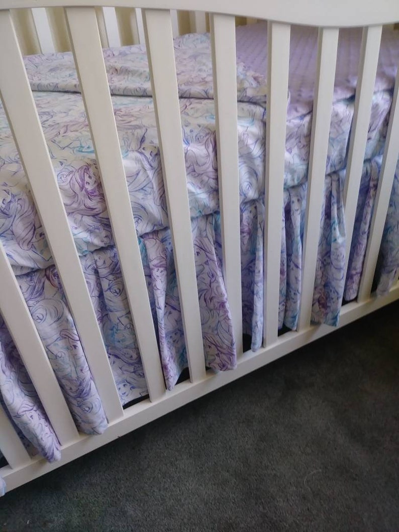 May include: White crib with a white and purple patterned fitted sheet and a white ruffled crib skirt. The crib skirt has a watercolor-like design of mermaids and seahorses.