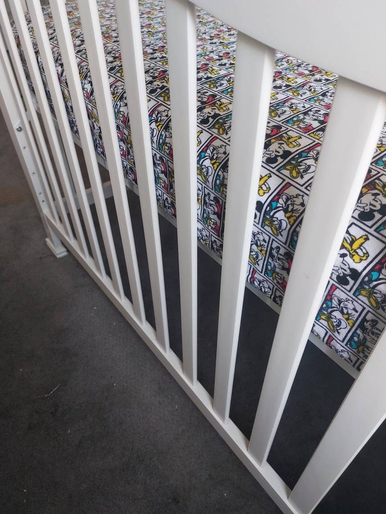 May include: White crib with white wooden slats and a fitted sheet with a cartoon character print. The sheet features a repeating pattern of Mickey Mouse, Donald Duck, Goofy, and Pluto.