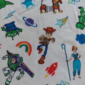 May include: White fabric featuring colorful cartoon characters from the Toy Story movie franchise. The design includes Woody, Buzz Lightyear, Slinky Dog, aliens, and other elements like stars, rainbows, and rockets.