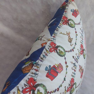 Peut inclure: Un oreiller blanc avec une bordure bleue. L'oreiller pr&eacute;sente un motif de No&euml;l festif avec un motif r&eacute;p&eacute;titif de couronnes vertes, de n&oelig;uds rouges et une ligne de personnes se tenant la main. Le texte "Joyeux No&euml;l !" est visible en bas de l'oreiller.