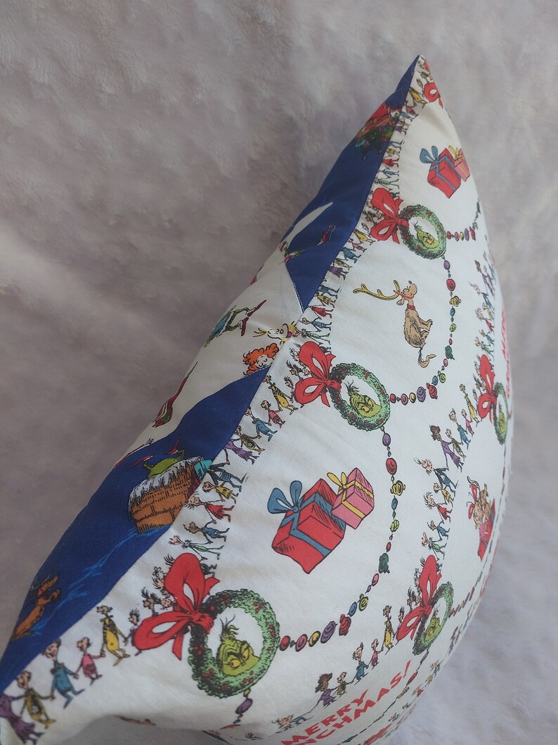 Peut inclure: Un oreiller blanc avec une bordure bleue. L'oreiller pr&eacute;sente un motif de No&euml;l festif avec un motif r&eacute;p&eacute;titif de couronnes vertes, de n&oelig;uds rouges et une ligne de personnes se tenant la main. Le texte "Joyeux No&euml;l !" est visible en bas de l'oreiller.