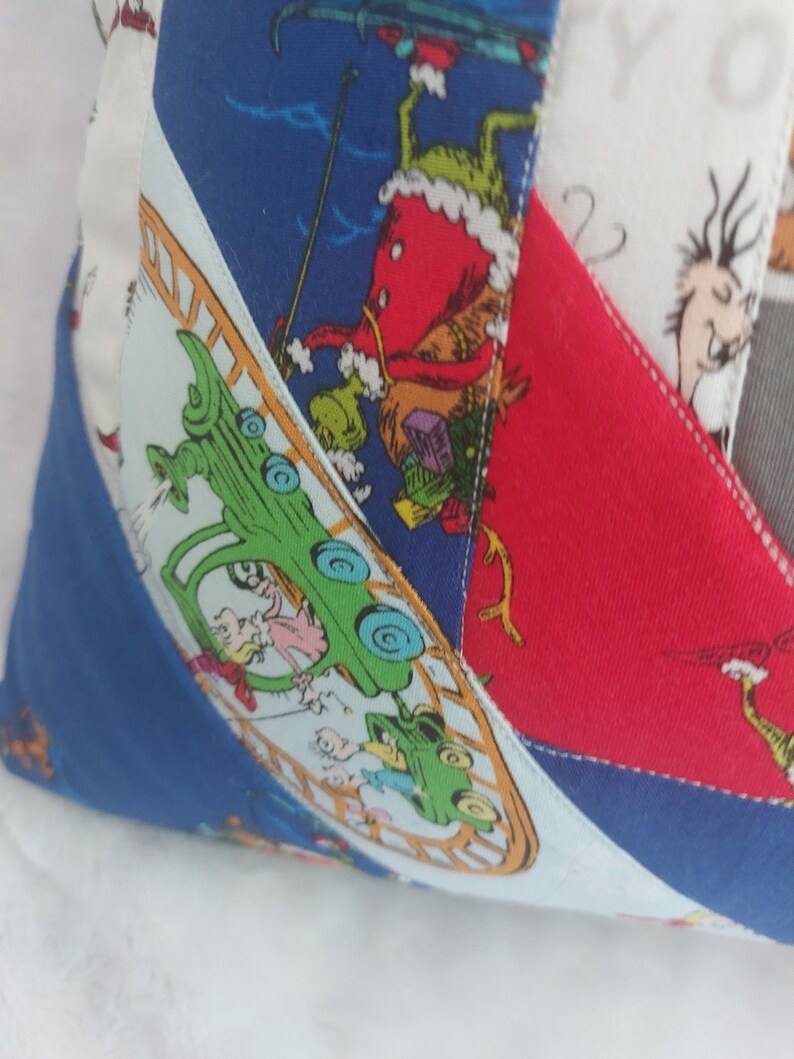 Peut inclure: Un oreiller patchwork color&eacute; avec un fond bleu, rouge et blanc. L'oreiller pr&eacute;sente une voiture verte avec des personnes &agrave; l'int&eacute;rieur, une figure rouge avec un chapeau vert et une figure blanche avec un chapeau noir. L'oreiller est fait de diff&eacute;rents tissus et a un design cousu.