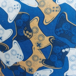 May include: A fabric with a repeating pattern of video game controllers in white, gold, and navy blue on a royal blue background. The controllers feature buttons and directional pads.