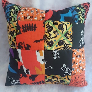 May include: A patchwork Halloween-themed pillow with a variety of colorful fabric squares. The squares feature orange, black, and yellow patterns with skeletons, bats, candy, and other spooky designs. The pillow has pointed corners.