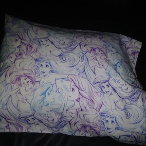 May include: A white pillowcase with a repeating pattern of purple and blue illustrations of Ariel, the Little Mermaid, with her long flowing hair.