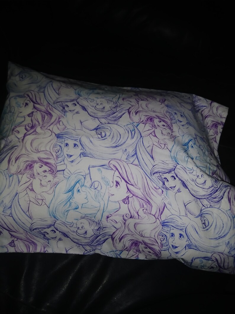 May include: A white pillowcase with a repeating pattern of purple and blue illustrations of Ariel, the Little Mermaid, with her long flowing hair.