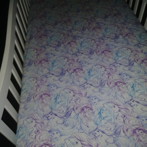 May include: A white crib sheet with a purple and blue pattern of Ariel, the Little Mermaid, repeated throughout the sheet.