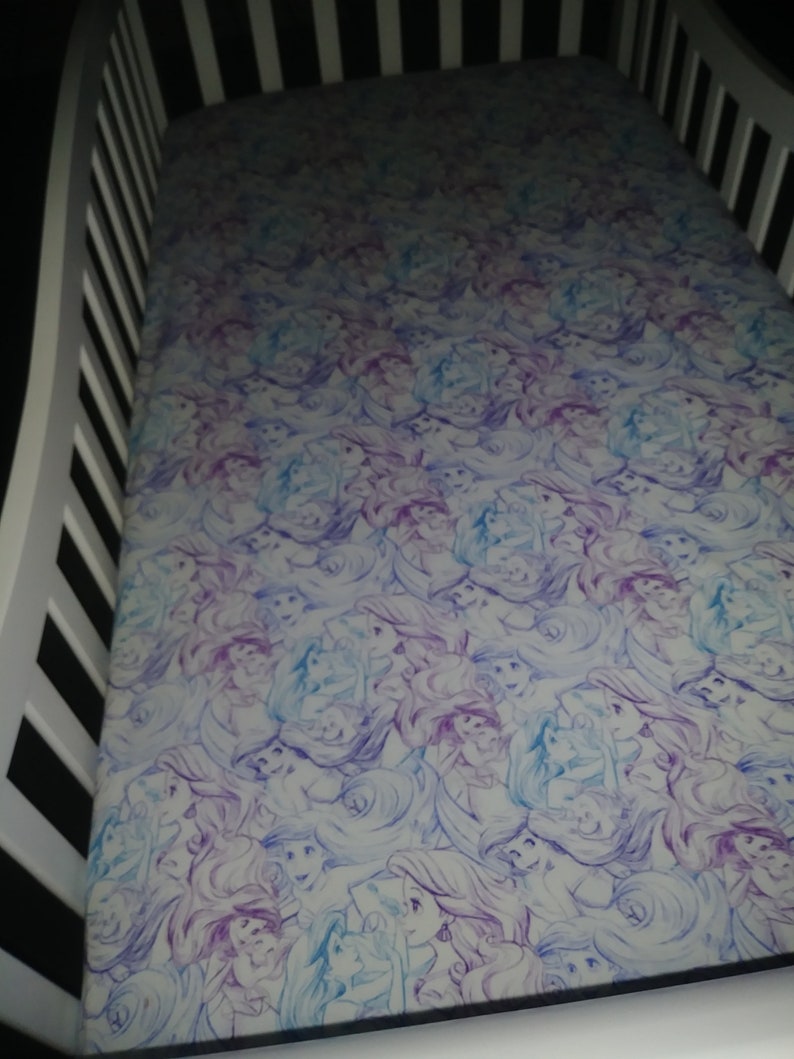 May include: A white crib sheet with a purple and blue pattern of Ariel, the Little Mermaid, repeated throughout the sheet.