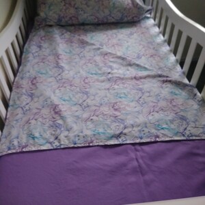 May include: A white crib with a purple fitted sheet and a white sheet with a purple and blue pattern. A white pillow with a purple and blue pattern is on top of the sheet.