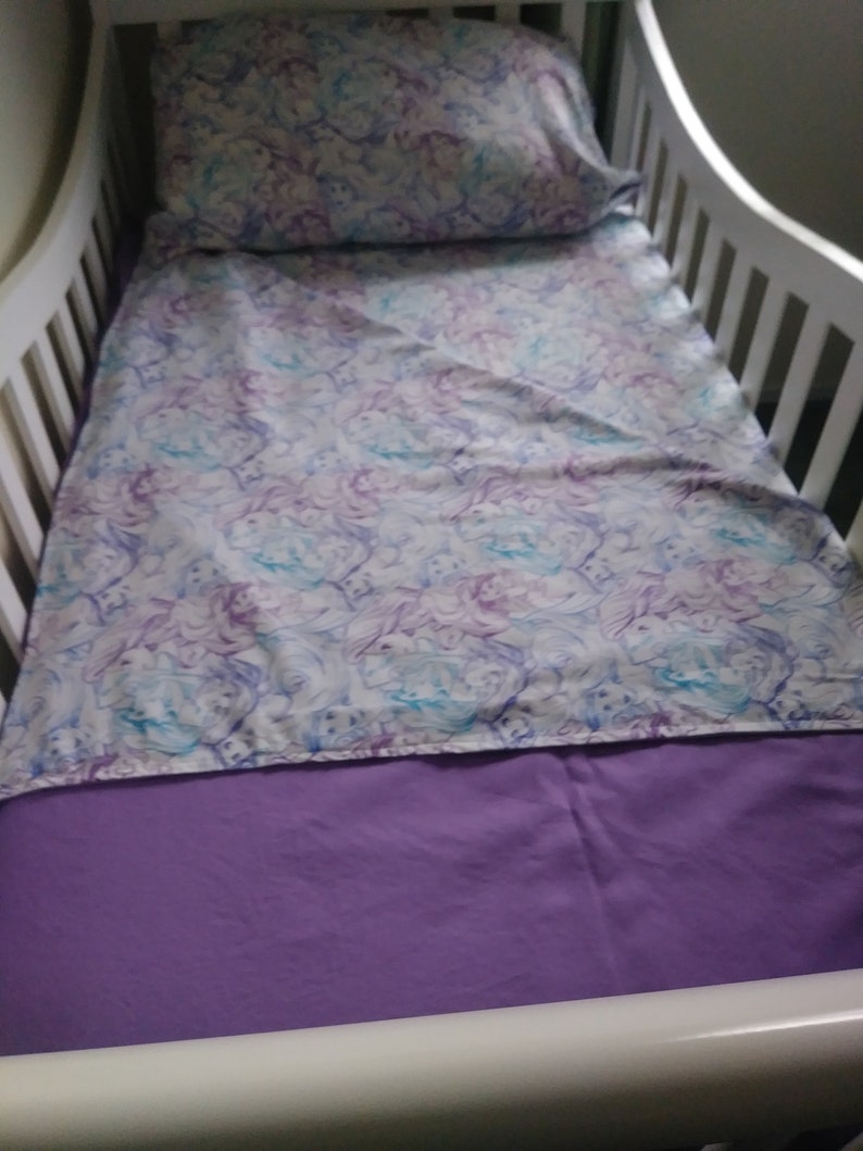 May include: A white crib with a purple fitted sheet and a white sheet with a purple and blue pattern. A white pillow with a purple and blue pattern is on top of the sheet.