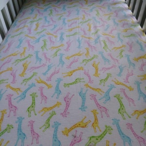 May include: White crib sheet with a repeating pattern of colorful giraffes in shades of pink, yellow, green, and blue. The giraffes are scattered across the sheet, creating a playful and whimsical design.