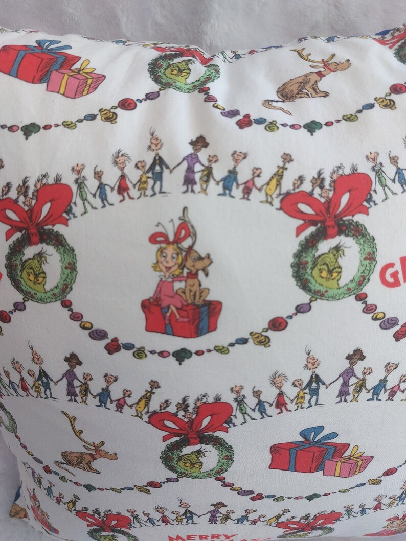 Peut inclure: Housse de coussin blanche avec un motif de No&euml;l festif repr&eacute;sentant le Grinch, un renne et un groupe d'enfants. Le motif comprend des couronnes, des cadeaux et les mots "Joyeux No&euml;l".
