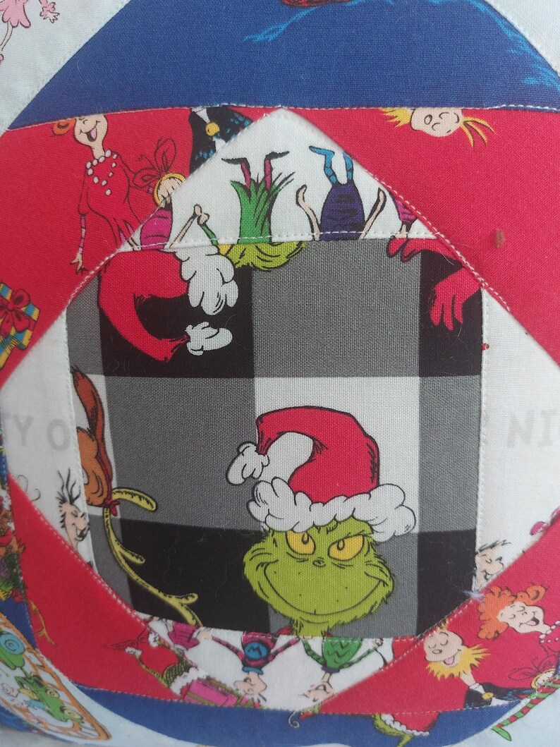 Peut inclure: Un tissu &agrave; carreaux rouge, blanc et noir avec un personnage vert du Grinch portant un chapeau de P&egrave;re No&euml;l rouge. Le Grinch est entour&eacute; d'autres carr&eacute;s de tissu color&eacute;s avec des personnages de dessins anim&eacute;s.
