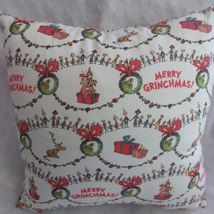 Peut inclure: Un oreiller blanc avec un motif r&eacute;p&eacute;t&eacute; d'une couronne verte, de n&oelig;uds rouges et d'un groupe de personnes se tenant la main. Le texte "Merry Grinchmas!" est r&eacute;p&eacute;t&eacute; tout au long du motif.