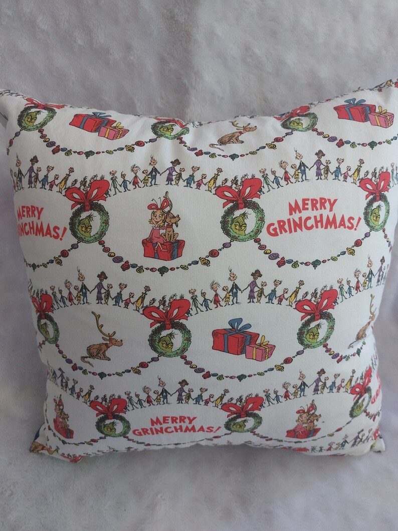 Peut inclure: Un oreiller blanc avec un motif r&eacute;p&eacute;t&eacute; d'une couronne verte, de n&oelig;uds rouges et d'un groupe de personnes se tenant la main. Le texte "Merry Grinchmas!" est r&eacute;p&eacute;t&eacute; tout au long du motif.