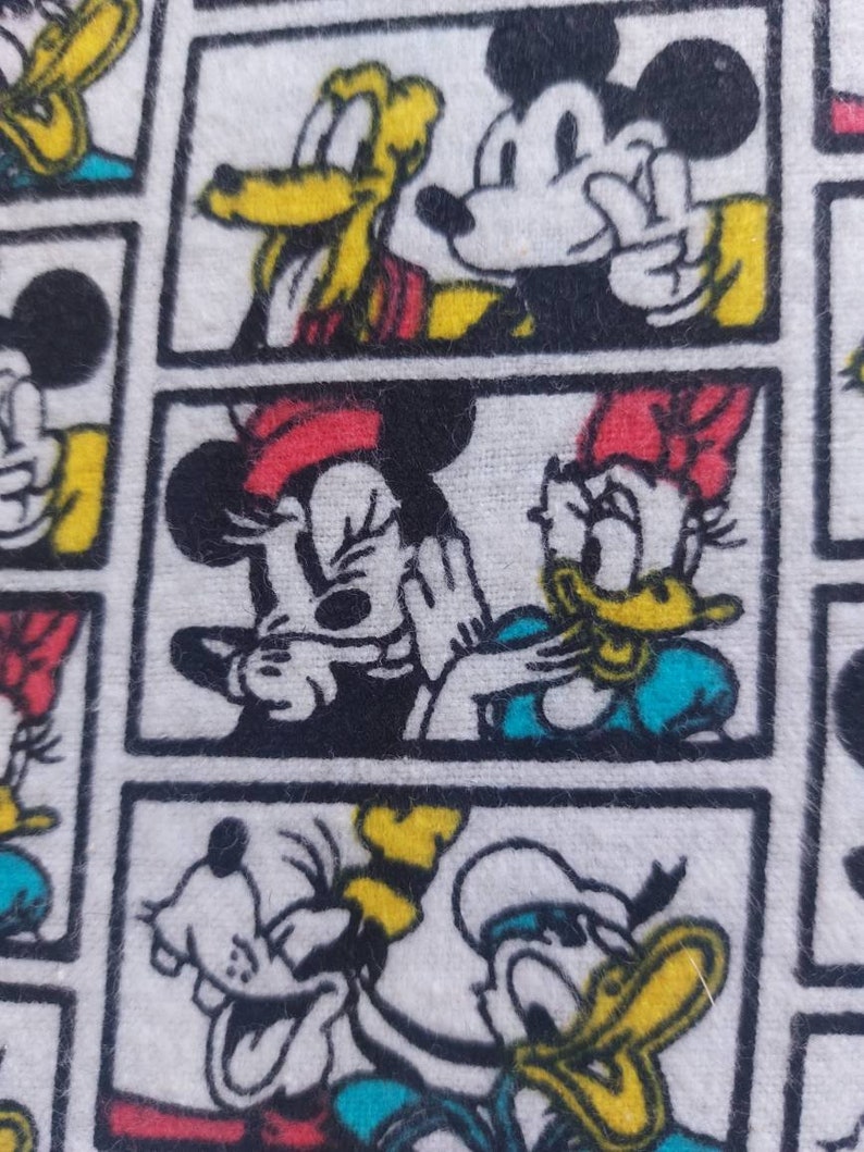 May include: A white fabric with a repeating pattern of cartoon characters. The characters include Mickey Mouse, Minnie Mouse, Donald Duck, and Goofy. The characters are in black, yellow, red, and blue.