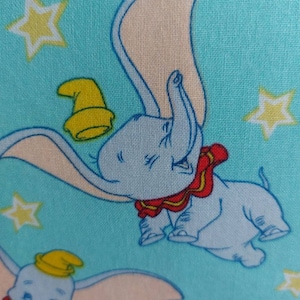 May include: A blue fabric with a repeating pattern of a cartoon elephant with a yellow hat and red and yellow trim. The elephant is flying with its trunk raised. The background is a light blue with yellow stars.