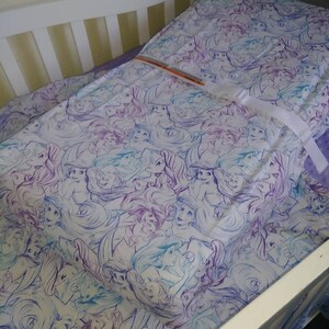 May include: A white crib with a purple and blue patterned fitted sheet featuring a repeated design of mermaids. The sheet is tucked into the crib and the crib is made of white wood.