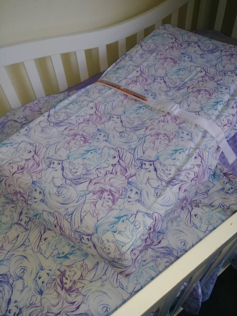 May include: A white crib with a purple and blue patterned fitted sheet featuring a repeated design of mermaids. The sheet is tucked into the crib and the crib is made of white wood.
