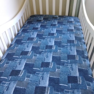 May include: A white crib with a blue denim-style patterned fitted sheet. The sheet features a patchwork design with various shades of blue and white, creating a textured appearance. The crib has a classic design with vertical slats.