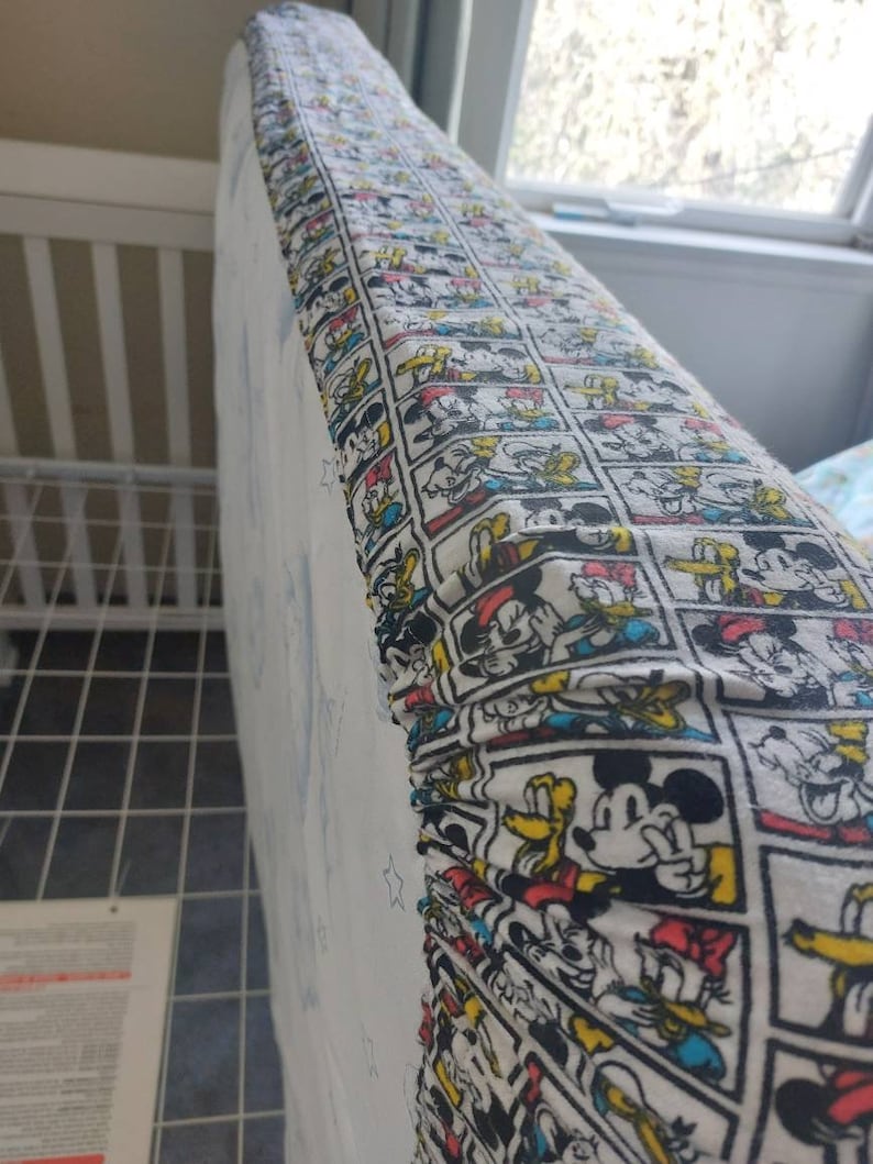 May include: A white fitted crib sheet with a colorful cartoon print featuring Mickey Mouse, Donald Duck, and Goofy. The print is repeated in rows across the sheet.