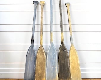 Könnte beinhalten: Fünf hölzerne Bootsruder, die aufrecht in einer Reihe stehen. Die Ruder haben ein abgenutztes Finish mit verschiedenen Farbkombinationen aus Grau, Dunkelgrau und metallischer Goldfarbe, die auf die Blätter und Griffe aufgetragen sind.