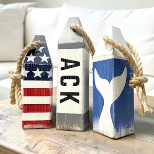 May include: Three wooden lobster buoy decorations with rope loops. The left buoy features a red, white, and blue American flag pattern. The center buoy is white and gray with the black text ACK. The right buoy is blue with a white whale tail.