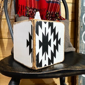 A white wooden tissue box cover with a black and white geometric pattern. The tissue box cover is sitting on a black wooden chair.