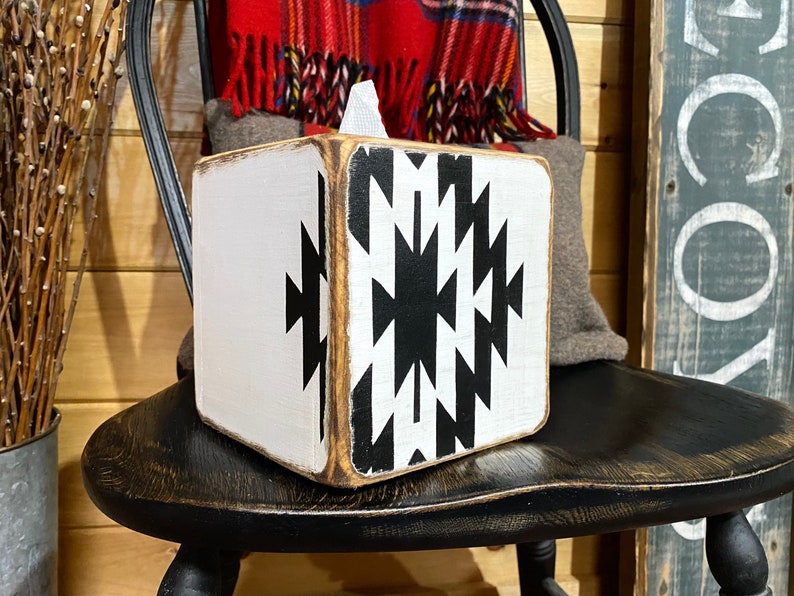 A white wooden tissue box cover with a black and white geometric pattern. The tissue box cover is sitting on a black wooden chair.