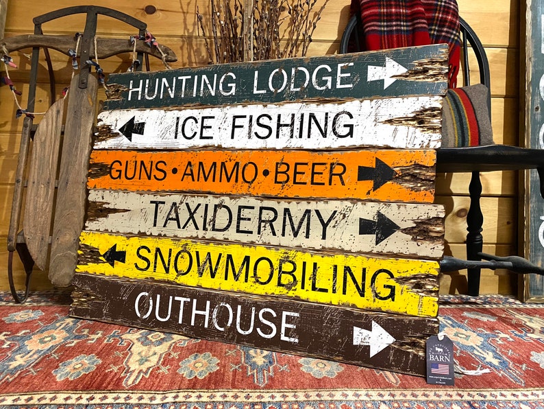 Puede incluir: Un letrero r&uacute;stico de madera con flechas direccionales que indica "Hunting Lodge", "Ice Fishing" y "Guns Ammo Beer". El letrero tiene un acabado envejecido con varios colores, incluyendo verde, blanco, naranja y amarillo, para una caba&ntilde;a.