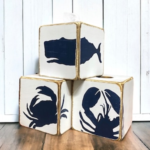 May include: Three white wooden tissue box covers with gold trim. Each box features a different navy blue silhouette: a whale, a crab, and a lobster.