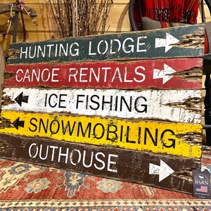 May include: A rustic wooden sign with five painted lines, each with a different activity and direction arrow. The activities are "Hunting Lodge", "Canoe Rentals", "Ice Fishing", "Snowmobiling", and "Outhouse".