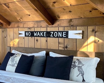 May include: A long, rectangular wooden sign with a distressed white finish and a central black horizontal stripe featuring the white text NO WAKE ZONE. The sign is mounted on a vertical wood-paneled wall above a bed with black and white pillows.