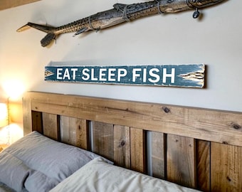 May include: A rectangular wooden wall sign with a distressed dark teal finish and white lettering that reads EAT SLEEP FISH. The sign features rough, weathered edges and is mounted horizontally on a wall above a wooden headboard.