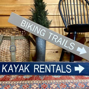 K&ouml;nnte beinhalten: Ein rechteckiges graues Holzschild mit wei&szlig;er Aufschrift BIKING TRAILS und einem Pfeil nach rechts, platziert &uuml;ber einem rechteckigen dunkelblauen Holzschild mit wei&szlig;er Aufschrift KAYAK RENTALS und einem Pfeil nach rechts.