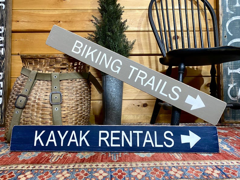K&ouml;nnte beinhalten: Ein rechteckiges graues Holzschild mit wei&szlig;er Aufschrift BIKING TRAILS und einem Pfeil nach rechts, platziert &uuml;ber einem rechteckigen dunkelblauen Holzschild mit wei&szlig;er Aufschrift KAYAK RENTALS und einem Pfeil nach rechts.