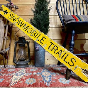 Rustic Snowmobile Trails Directional Sign displayed in a cozy log cabin, hand-painted and distressed for a weathered, vintage look. Perfect for mountain cabins, winter vacation homes, ski lodges, or rustic retreats, this handcrafted sign adds northern trail charm and rustic adventure style. Handmade in Maine with artisan craftsmanship