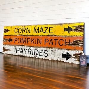 May include: A weathered wooden sign with three rows of text, each with a black arrow pointing to the right. The top row reads "CORN MAZE", the middle row reads "PUMPKIN PATCH", and the bottom row reads "HAY RIDES".