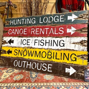 May include: A rustic wooden sign with five painted planks, each with a different activity and direction arrow. The top plank reads "Hunting Lodge", the second plank reads "Canoe Rentals", the third plank reads "Ice Fishing", the fourth plank reads "Snowmobiling", and the bottom plank reads "Outhouse".