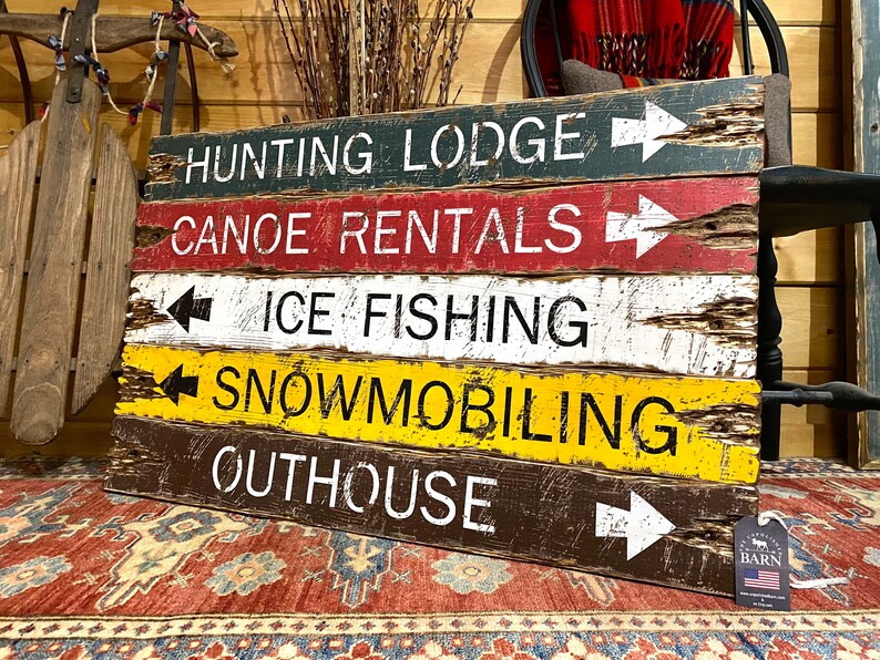 May include: A rustic wooden sign with five painted planks, each with a different activity and direction arrow. The top plank reads "Hunting Lodge", the second plank reads "Canoe Rentals", the third plank reads "Ice Fishing", the fourth plank reads "Snowmobiling", and the bottom plank reads "Outhouse".
