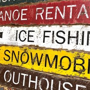 May include: A weathered wooden sign with four rows of text. The top row says "Canoe Rentals". The second row says "Ice Fishing" with a left-pointing arrow. The third row says "Snowmobil" with a left-pointing arrow. The bottom row says "Outhouse".