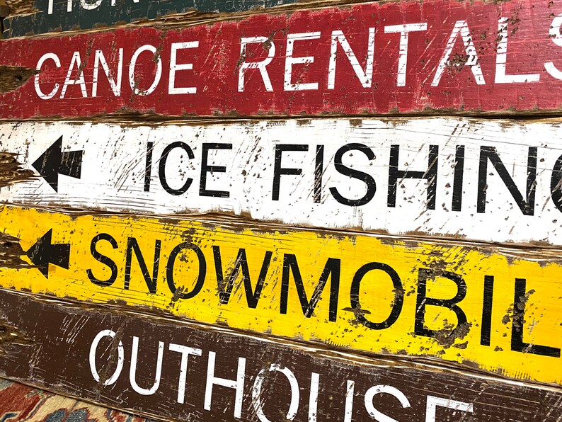May include: A weathered wooden sign with four rows of text. The top row says "Canoe Rentals". The second row says "Ice Fishing" with a left-pointing arrow. The third row says "Snowmobil" with a left-pointing arrow. The bottom row says "Outhouse".